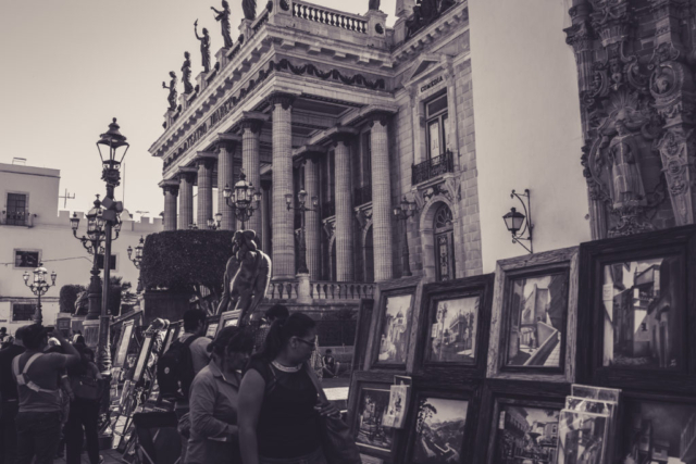 Black and white photo of an art exhibit in the street of Irapuato, in Mexico. In the background, there is the opera and in the foreground people looking at the paintings.