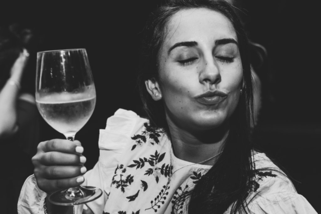 Black and white photo of a young woman closing her eyes and toasting with a white wine glass in her left hand.