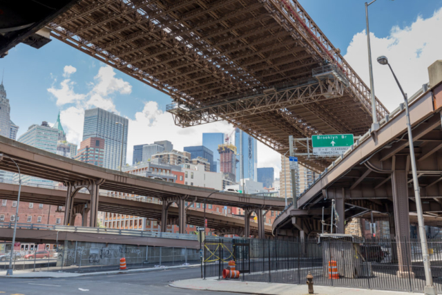 Photo of a lot of bridges in New York. In the background we can see NY downtown with its huge buildings and in the foreground, bridges and green road signs showing the direction of Brooklyn Bridge.