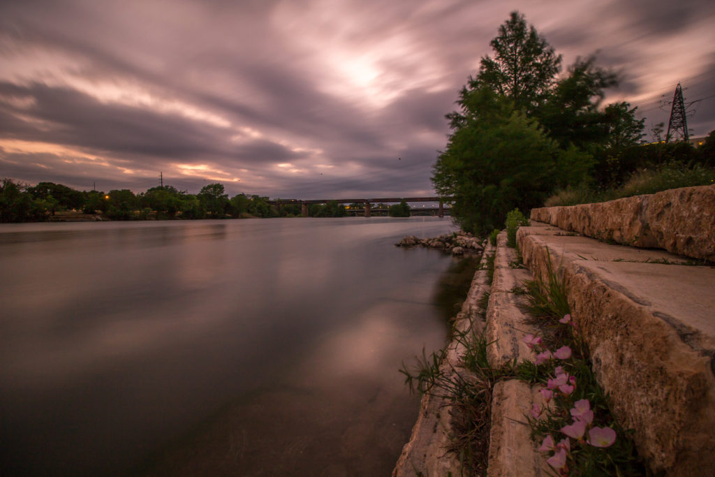 Sunset of Town Lake in Austin, Texas . In the foreground, there are rocks and in the background a bridge. Clouds are heavy and low.