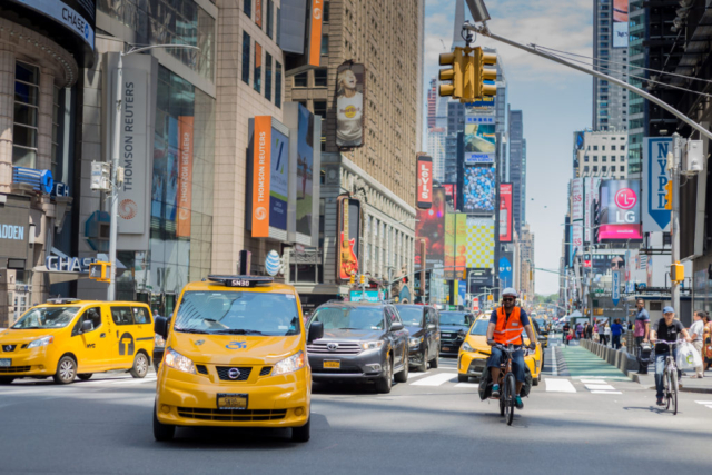 Photo of a New York street next to Time Square. People riding, walking on the sidewalk. Cars and taxis driving are about to turn right. Huge buildings with commercials promoting LG, Chase,...