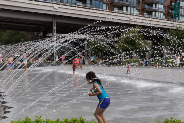 Photo of kids playing in the water in a parc in Georgetown - Washington DC. In the foreground, a girl wearing a blue tee-shirt and short is running and playing with the water.